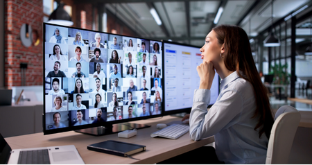 Projektleiterin stützt das Kinn auf die Hand und beobachtet ein virtuelles Meeting mit großem Team am Monitor.