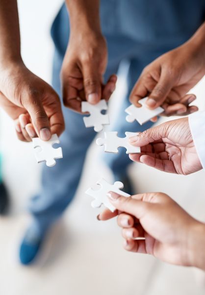 Nahaufnahme mehrerer Hände mit weißen Puzzleteilen über einem hellen Büroboden. 